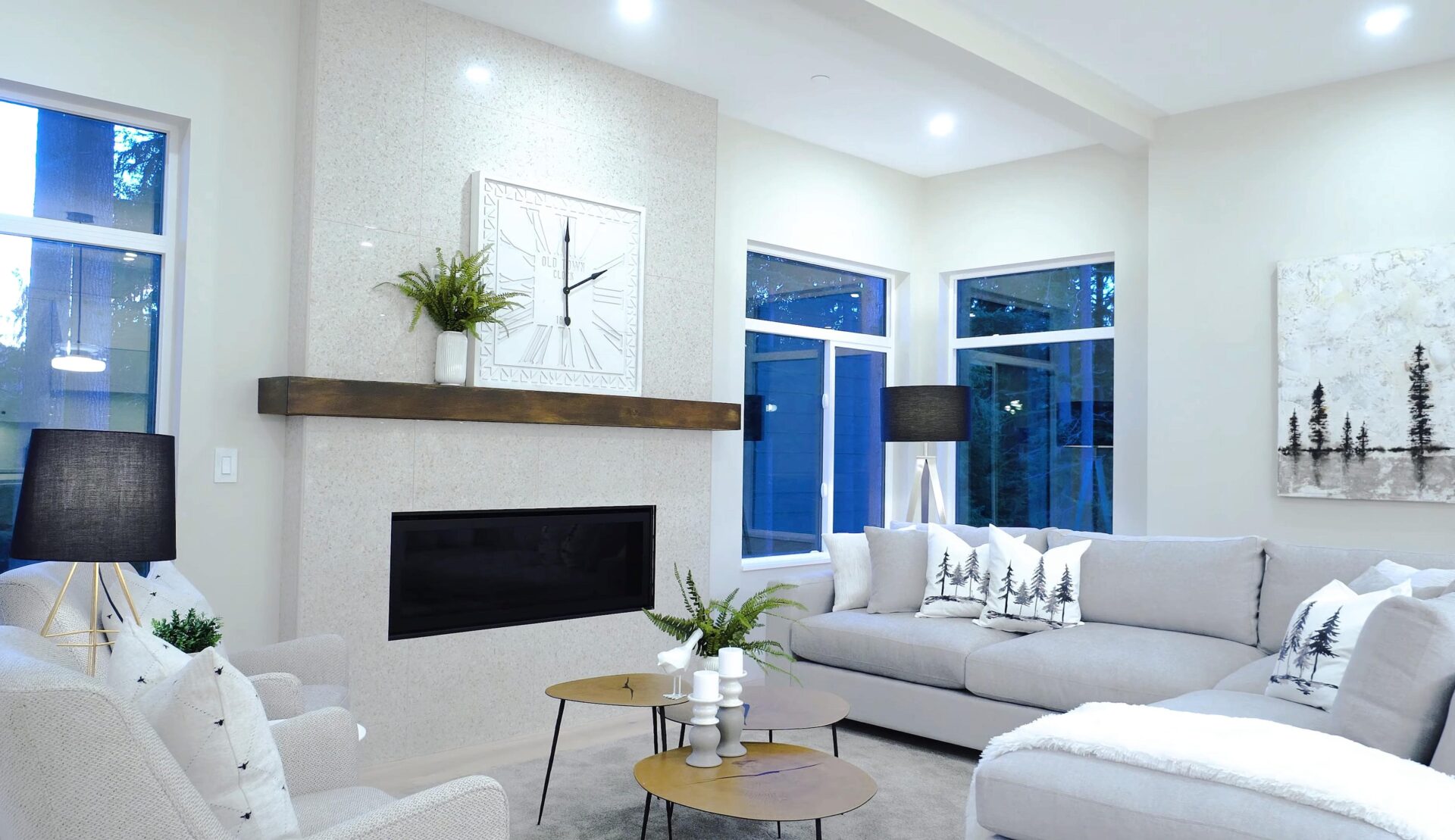 shutterstock_1925921891-min A sleek white living room centered around a modern fireplace with a farmhouse-style wooden mantel. There is a large square clock on the mantel set to two o'clock and a small fern plant beside it. A sectional sofa and two armchairs are arranged around three round wooden coffee tables of varying heights. Several throw pillows on the sectional depict artfully rendered evergreen trees, as does a painting on the wall behind the sectional.